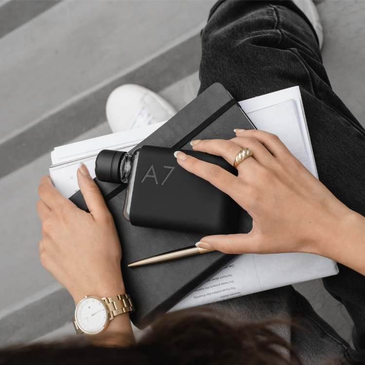 Woman holding a black notebook with 'A7' branding on a 180ml water bottle, wearing a gold watch and ring.