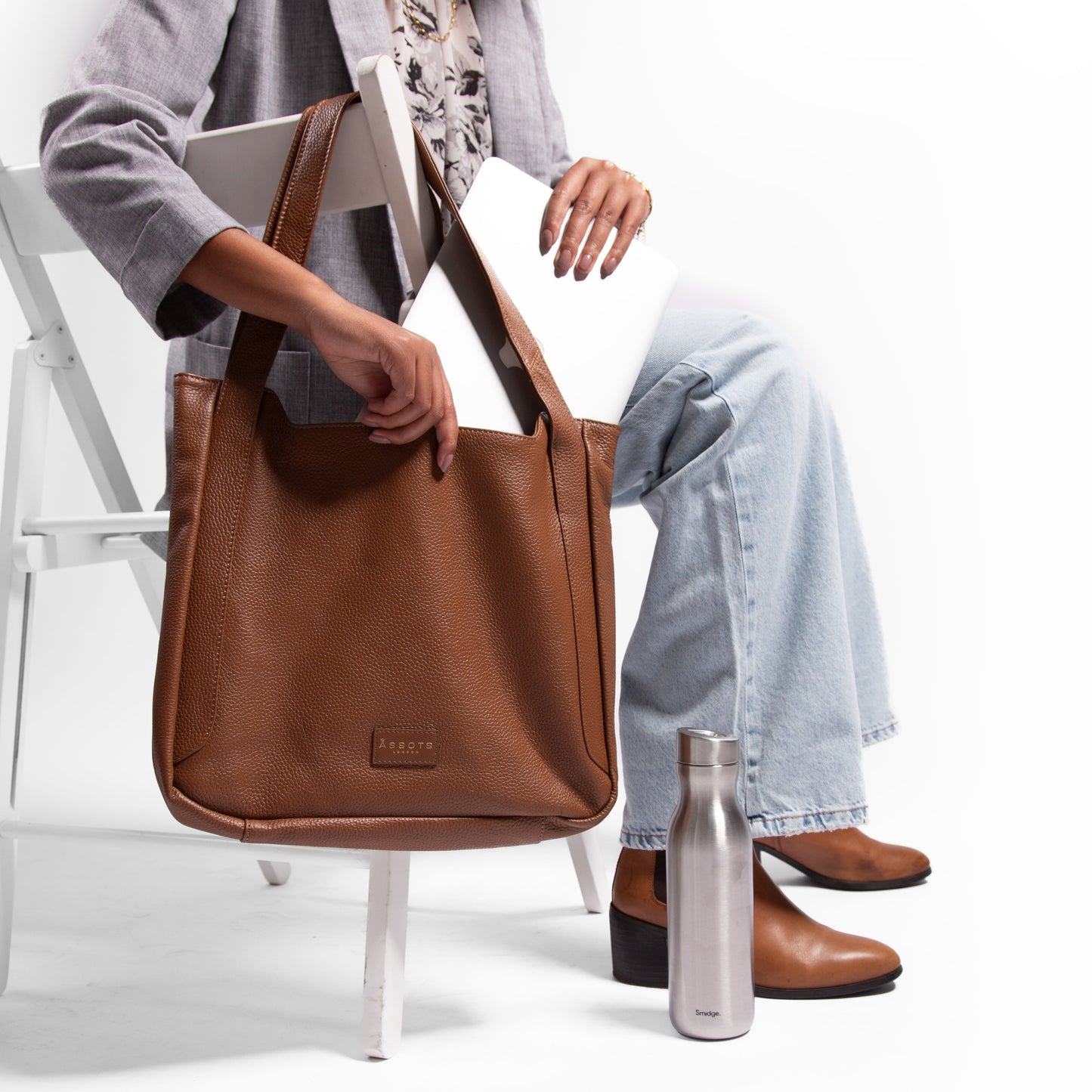 Woman sitting on a chair with a brown leather handbag putting a laptop inside it and a silver stainless steel 500ml water bottle on the ground 